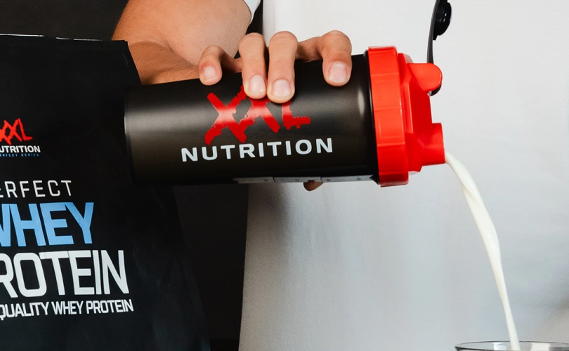 Hand schenkt een eiwitshake uit een shaker in een glas met op de shaker het logo van XXL Nutrition.